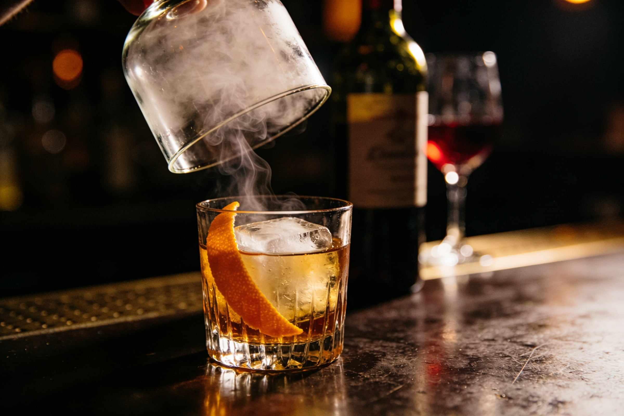 An old fashioned cocktail with orange peel on a bar, smoke rising as a glass cloche is lifted