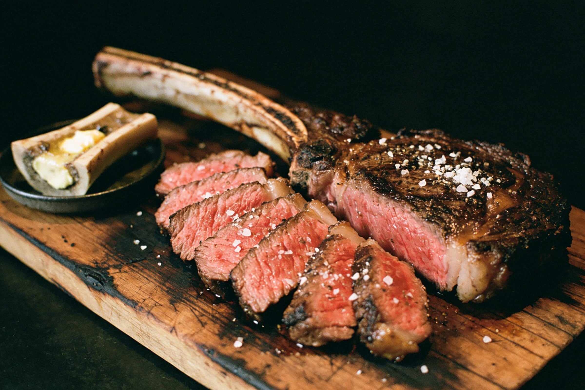 A sliced bone-in ribeye, pink inside with a dark crust and flakes of salt, on a scorched oak board beside a roasted marrow bone with butter