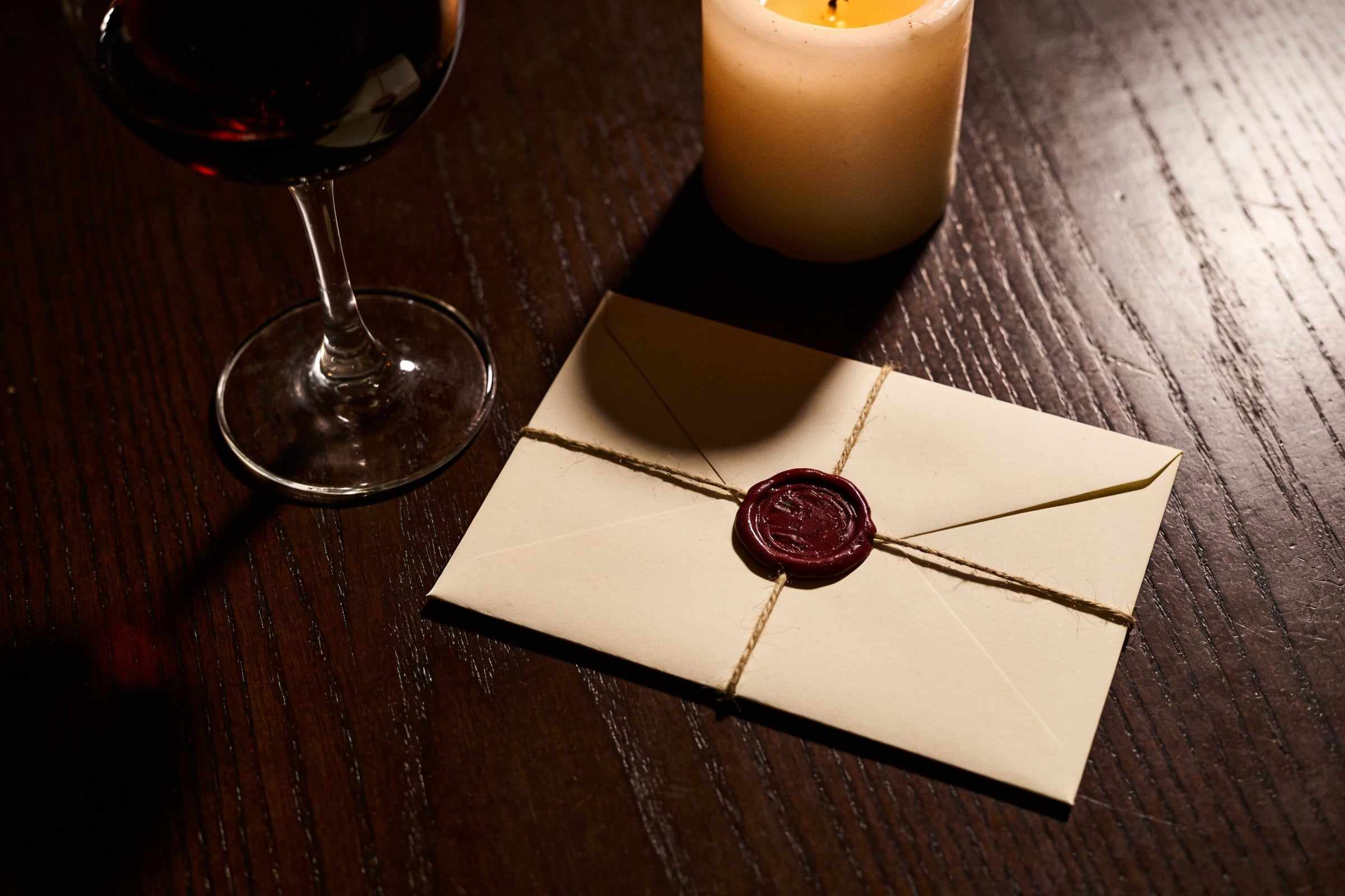 A cream envelope sealed with dark red wax and tied with string, beside a glass of red wine and a candle