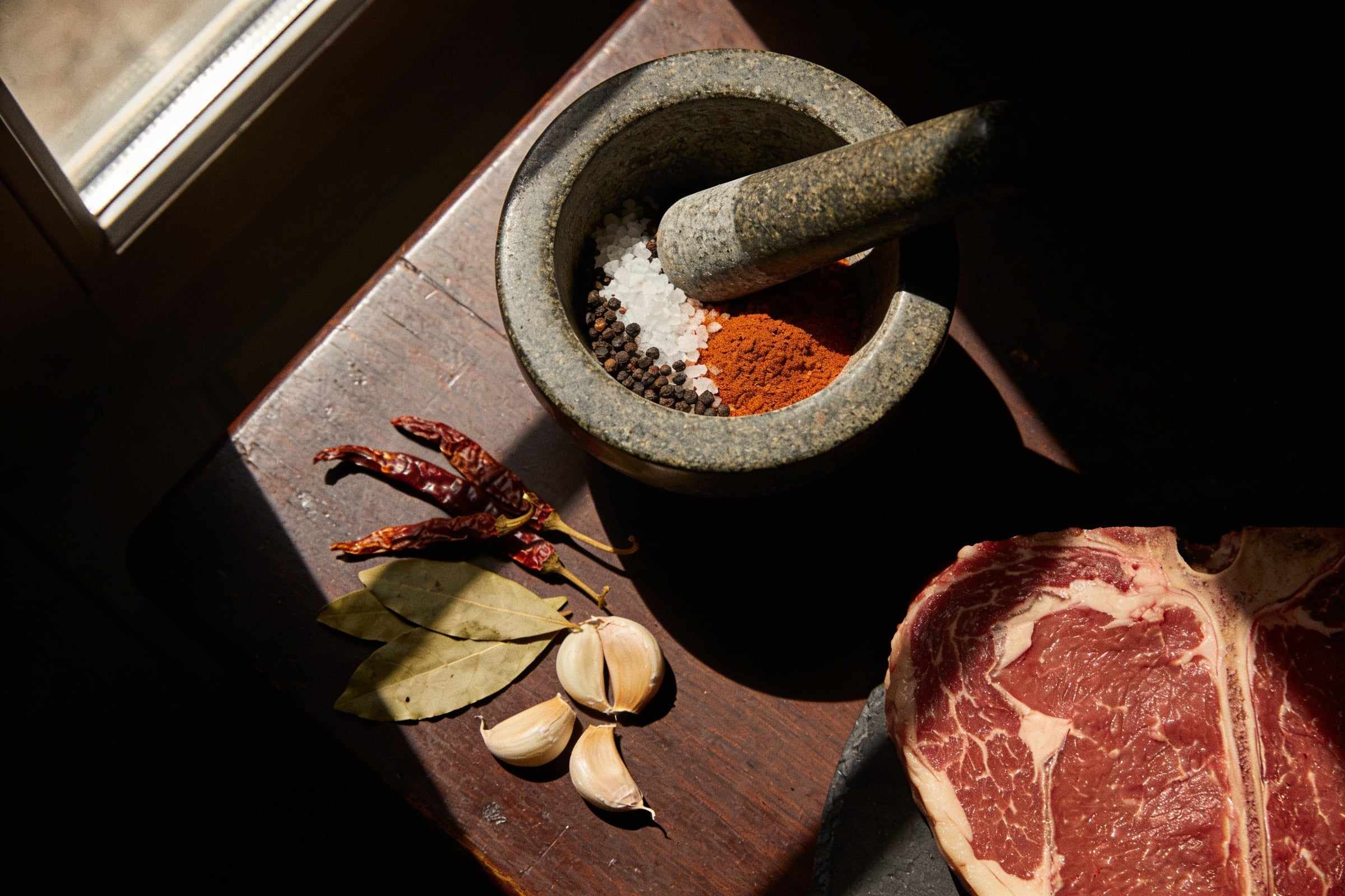 A stone mortar with black pepper, sea salt and smoked paprika on a dark table in a shaft of light, a raw steak at the edge