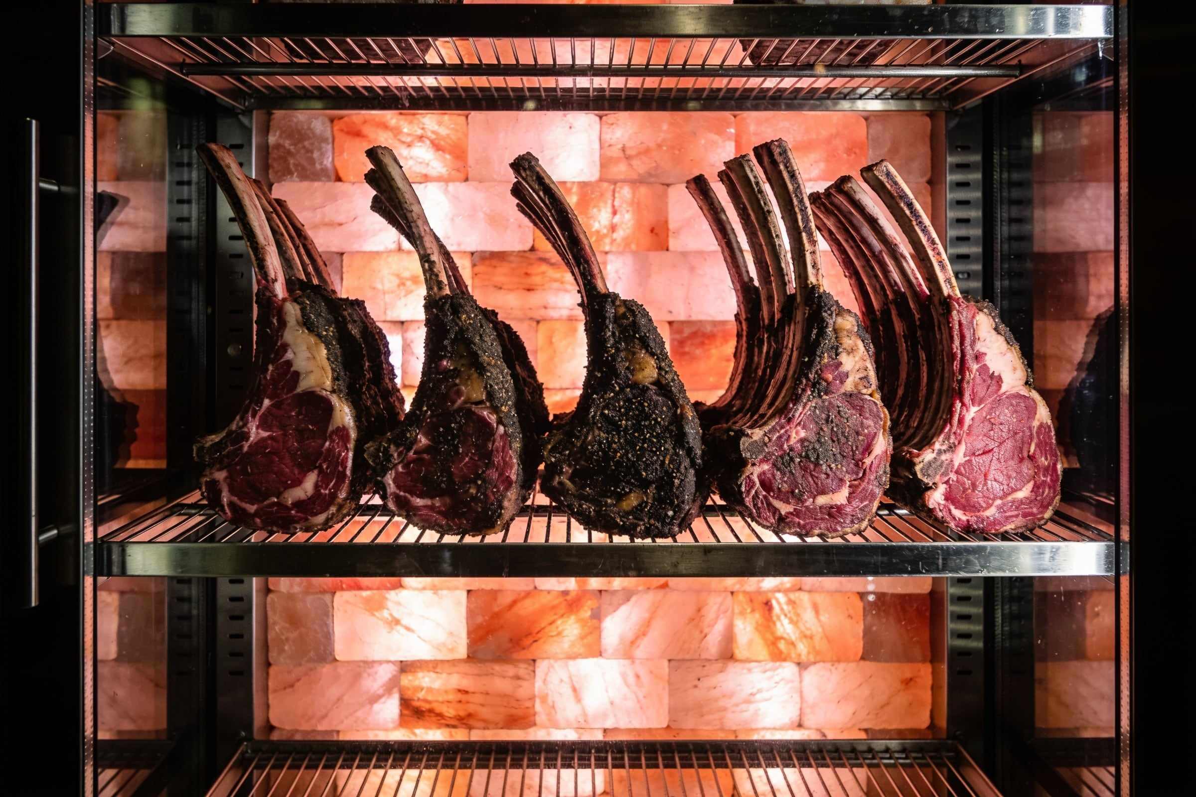 Racks of beef ribs with a dark aged crust inside a glass cabinet, a wall of glowing pink salt blocks behind them