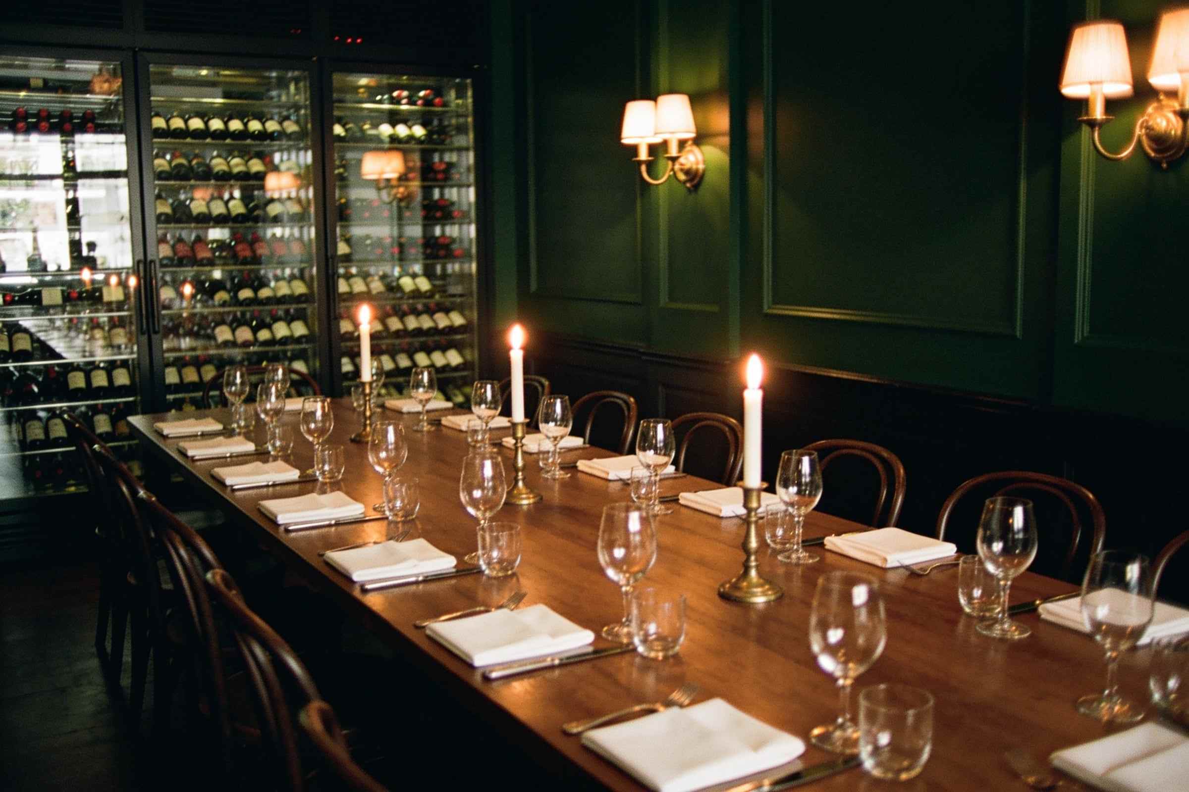 A long table set for fourteen with candles and white napkins in a dark green room lit by brass wall lamps